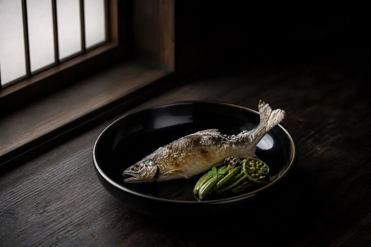 黒漆の器に盛られた川魚の塩焼きと旬の山菜。窓からの自然光がひと筋差す(イメージ)